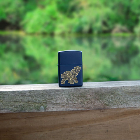 Lifestyle image of Lucky Elephant Design Navy Matte Windproof Lighter standing on a rail, with a pond and trees in the background.