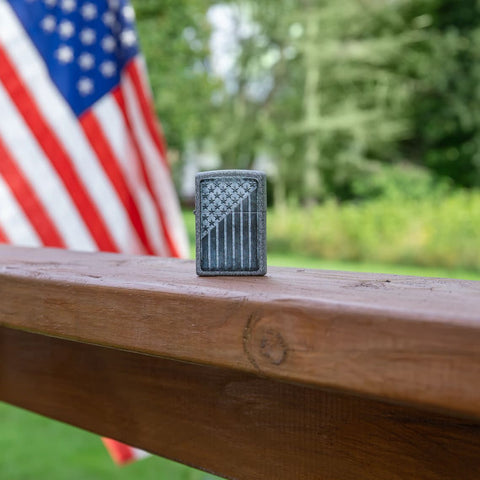 Lifestyle image of Stars and Stripes Design Iron Stone Windproof Lighter standing on a railing with an American Flag in the background.
